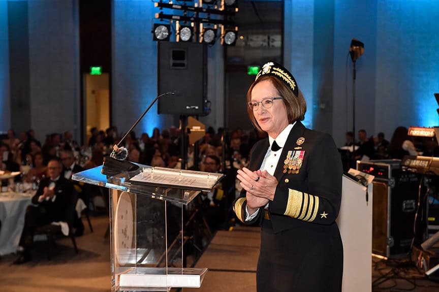 A female US Navy officer in dress uniform stands at a transparent podium, gesturing with her hands while speaking at a formal banquet event
