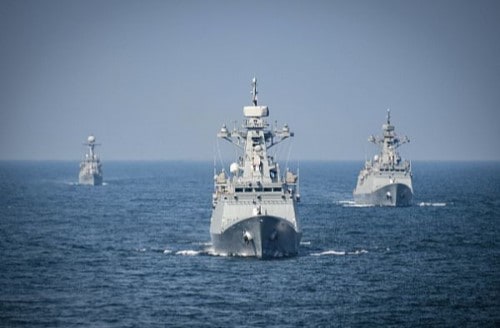 Three U.S. Naval vessels traveling over calm seas directly toward the camera.