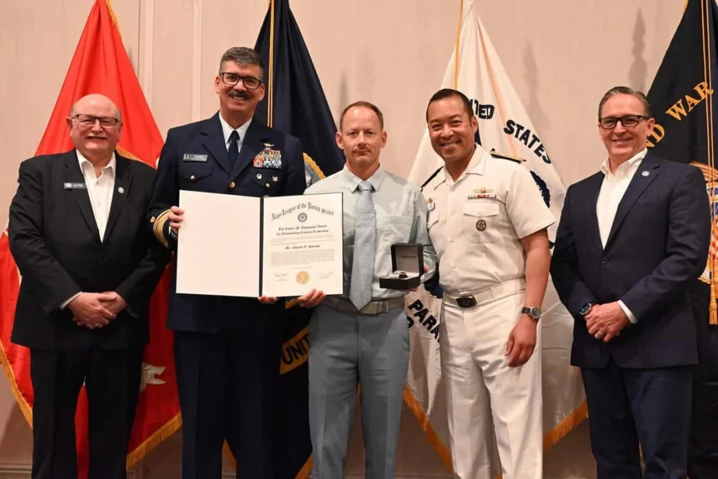 5 men stand in a row with flags behind them. One is holding and award.